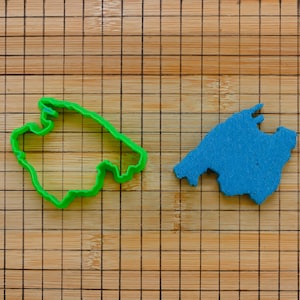 Puede incluir: Un cortador de galletas de plástico verde con forma de contorno de mapa se encuentra junto a un recorte de fieltro azul de un mapa. Ambos están sobre una tabla de cortar de madera.