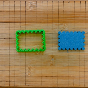 Può includere: Un tagliapasta di plastica verde a forma di rettangolo con bordi smerlati e un biscotto di schiuma blu a forma di rettangolo con bordi smerlati su un tagliere di legno.
