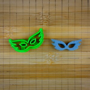 May include: Two plastic cookie cutters shaped like masquerade masks. The mask on the left is green and the mask on the right is blue.