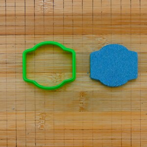 Può includere: Un tagliapasta di plastica verde e un ritaglio di pasta di zucchero blu, entrambi a forma di rettangolo con angoli arrotondati, su un tagliere di legno.