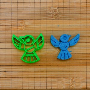 Puede incluir: Un cortador de galletas de plástico verde brillante con forma de pájaro junto a una figura de pájaro azul hecha de material granular. El diseño del pájaro presenta alas extendidas, una cabeza circular y una cola en abanico con detalles.