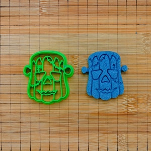 Può includere: Un tagliabiscotti in plastica verde brillante a forma di volto di mostro con bulloni sui lati e una sagoma in pasta blu testurizzata abbinata con lo stesso design, caratterizzato da dettagli di cuciture sulla fronte e una bocca a zig-zag.