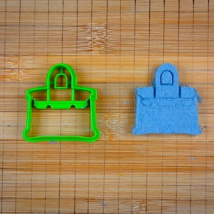 Op de afbeelding: Een groene plastic koekjesvormpje en een blauwe suikerkoek, beide in de vorm van een handtas.
