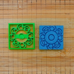 May include: A bright green square cookie cutter featuring a central circular design with radiating curved shapes and a matching blue square stamp with the same intricate circular pattern, both designed for creating detailed impressions in dough.