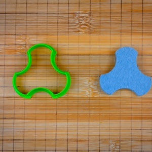 May include: A green plastic cookie cutter and a blue felt tri-star shape on a wooden cutting board.