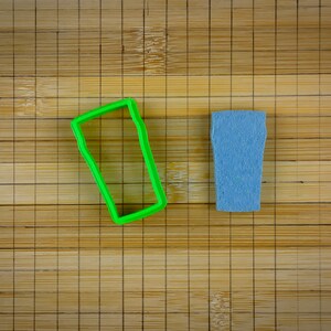 Op de afbeelding: Een groene plastic koekjesvormpje in de vorm van een glas bier naast een blauw glas bier op een bamboe snijplank.
