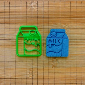May include: A bright green plastic milk carton cookie cutter next to a textured blue milk carton shape. Both items feature a graphic design of a milk carton with the word MILK embossed on the front, showing a pouring liquid detail.