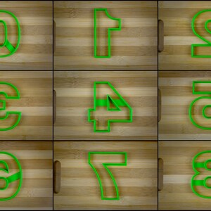 Puede incluir: Nueve cortadores de galletas de plástico verde en forma de los números del 0 al 8. Los cortadores están dispuestos en tres filas de tres sobre una superficie de madera.