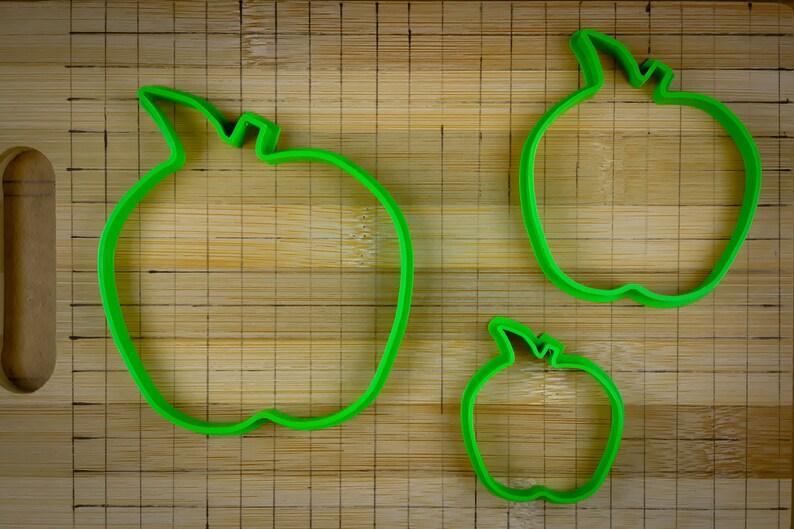 Peut inclure: Trois emportes-pi&egrave;ces en forme de pomme en plastique vert de diff&eacute;rentes tailles sur une planche &agrave; d&eacute;couper en bois.