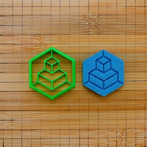 May include: A bright green hexagonal cookie cutter featuring an internal geometric cube design alongside a matching blue hexagonal piece of textured material with the same embossed cube pattern, both resting on a wooden surface with grid lines.