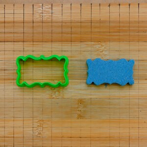 May include: A green plastic cookie cutter and a blue sponge, both shaped like a rectangle with wavy edges, are on a wooden cutting board.