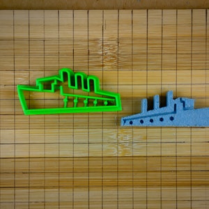 Può includere: Due stampini per biscotti a forma di nave, uno verde e uno blu, su un tagliere di bambù.