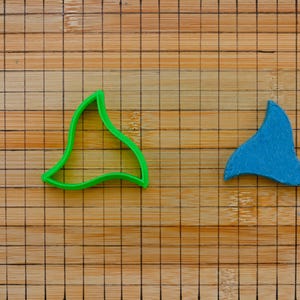 Può includere: Un tagliabiscotti in plastica verde e un ritaglio in feltro blu, entrambi a forma di coda di balena, sono esposti su un tagliere di legno. Il tagliabiscotti è di un verde brillante, mentre la coda in feltro è blu tinta unita.