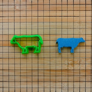 Op de afbeelding: Een groene, schaapvormige koekjesvorm en een blauw vilten schaap worden getoond op een bamboe snijplank. De koekjesvorm is een heldergroene omtrek, terwijl het vilten schaap een effen blauwe vorm is.