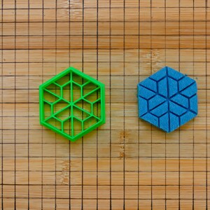 Könnte beinhalten: Zwei sechseckige Objekte, eines in leuchtendem Grün und eines in Blau, auf einer Holzoberfläche. Das grüne Objekt hat ein offenes, gitterartiges Design, während das blaue ein festes geometrisches Muster hat. Die Holzoberfläche ist kariert.