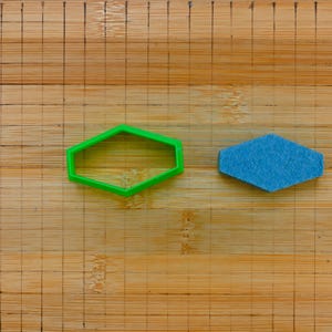 May include: A green plastic geometric shape and a blue felt geometric shape are displayed on a wooden cutting board. The cutting board has a grid pattern and a rounded cutout on the left side.