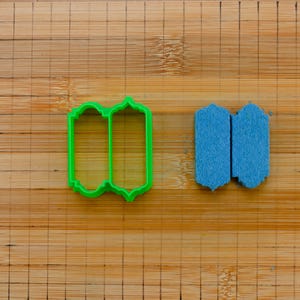 May include: Two cookie cutters on a wooden surface. The cutter on the left is green and shaped like a double-sided rectangle with rounded corners. The cutter on the right is blue and shaped like a double-sided rectangle with pointed corners.