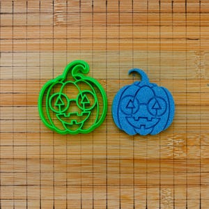 Op de afbeelding: Een felgroene koekjesvorm in de vorm van een pompoen naast een blauwe koekjesafdruk in de vorm van een pompoen. Beide items hebben een uitgesneden gezicht met een ronde bril, driehoekige ogen en een gekarteld mondontwerp.