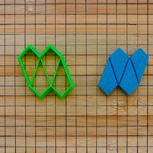 May include: Two geometric cookie cutters, one green and one blue, on a wooden surface. The green cutter is shaped like a diamond with multiple points, while the blue cutter is shaped like a letter W.