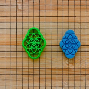 Op de afbeelding: Twee koekjesvormen, een groene en een blauwe, in de vorm van een levensbloemontwerp. De koekjesvormen liggen op een houten snijplank.