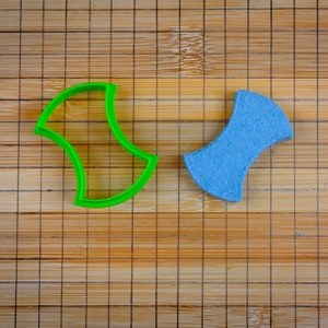 May include: A green plastic cookie cutter and a blue cookie in the shape of a bow tie on a bamboo cutting board.