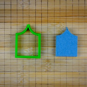 May include: A green plastic cookie cutter shaped like a house with a pointed roof sits next to a blue felt house cutout on a bamboo cutting board.