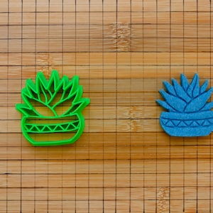 Könnte beinhalten: Zwei Ausstecher in Form von Sukkulenten. Einer ist leuchtend grün, der andere blau. Beide haben ein detailliertes Pflanzendesign in einem dekorativen Topf. Die Ausstecher liegen auf einem Bambus-Schneidebrett.