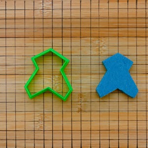 Op de afbeelding: Een groene plastic koekjesvorm en een blauwe vilten vorm liggen op een houten snijplank. De koekjesvorm heeft de vorm van een persoon. De vilten vorm heeft een effen blauwe kleur.