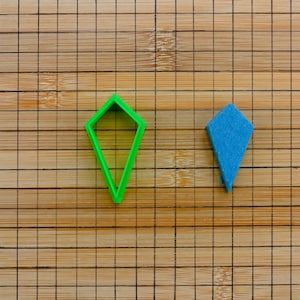 Könnte beinhalten: Zwei Ausstecher auf einer Holzoberfläche. Der Ausstecher links ist grün und diamantförmig. Der Ausstecher rechts ist blau und tropfenförmig.