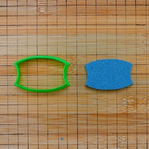Puede incluir: Un cortador de galletas de plástico verde y un objeto azul texturizado están sobre una tabla de cortar de bambú. El cortador de galletas tiene forma ovalada con lados curvos. El objeto azul es de forma sólida con superficie texturizada.