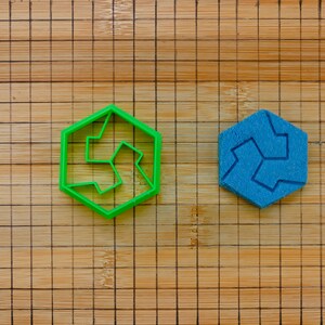 Może przedstawiać: Dwa sześciokątne elementy układanki, jeden zielony i jeden niebieski, leżą na drewnianej desce do krojenia z wzorem kratki. Zielony element to plastikowy kontur, a niebieski to pełny kształt. Elementy są zaprojektowane tak, aby pasowały do siebie.