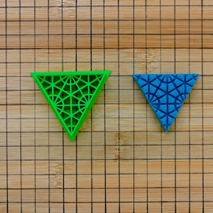 May include: Two geometric triangle shapes, one green and one blue, with a patterned design. The triangles are on a wooden surface with a grid pattern.