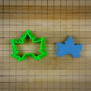May include: A green plastic maple leaf cookie cutter and a blue felt maple leaf on a wooden cutting board.