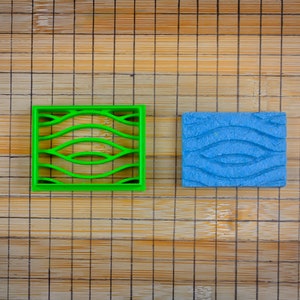 Può includere: Due stampini per biscotti, uno verde e uno blu, con un motivo ondulato. Gli stampini sono su un tagliere di bambù.