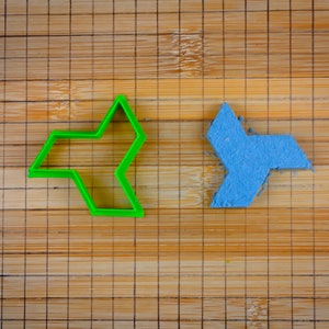 May include: Two plastic cookie cutters on a bamboo chopping board. The cutter on the left is green and shaped like a four-pointed star. The cutter on the right is blue and shaped like a three-pointed star.