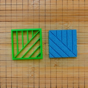 May include: A square green plastic cookie cutter featuring a geometric pattern of parallel lines and a matching blue square textured stamp with the same diagonal line design, both resting on a wooden surface with a printed grid pattern.