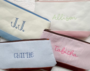 May include: Four personalized cosmetic bags with different patterns and colors. The bags are white with blue, pink, and brown accents. The names "J.J.", "Allison", "Charlie", and "Tabitha" are embroidered on the bags.