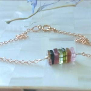 Puede incluir: Un delicado brazalete de cadena dorada con un dije de piedra preciosa multicolor. El dije está hecho de piedras facetadas apiladas en tonos de rosa, verde y negro.