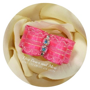 May include: A pink dog bow with gold glitter and two light blue rhinestones. The bow is on a white background.