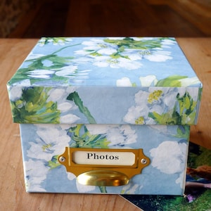 May include: A square photo storage box with a floral pattern in blue, white, and green. The box features a brass label holder with the word "Photos". A photograph is partially visible beside the box.