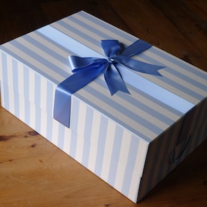 Puede incluir: Una caja de regalo grande y rectangular con un patrón de rayas azules y blancas. La caja está atada con una cinta de raso azul y un lazo.