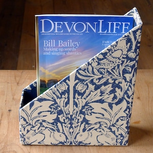 May include: A blue and white magazine holder with a floral pattern. The magazine holder is made of cardboard and has a decorative design. The magazine holder is holding a magazine titled "Devon Life" with the text "Bill Bailey Making up words and singing shanties".