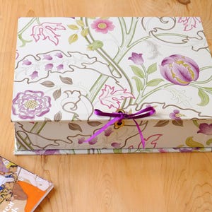Op de afbeelding: Een rechthoekige doos met een bloemenpatroon van paarse en roze bloemen, groene bladeren en een paars lint. De doos staat op een houten oppervlak, met een tijdschrift zichtbaar in de linker benedenhoek.