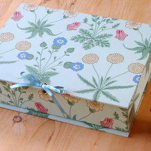 May include: A rectangular box featuring a floral design in green, pink, and yellow on a pale blue backdrop. It's secured with a light blue ribbon. A small vase with pink flowers is also visible.