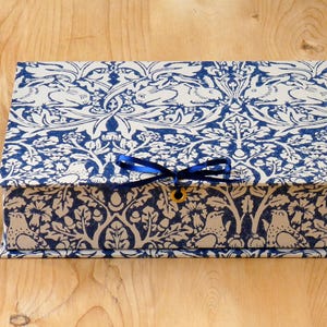 Könnte beinhalten: Eine rechteckige Box mit blau-weißem Blumenmuster. Die Box ist mit einem blauen Band und einem goldfarbenen Verschluss verschlossen. Links von der Box befindet sich eine kleine Vase mit rosa Blumen.