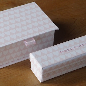 Puede incluir: Dos cajas rosas con un patrón de corazones blancos. La caja más pequeña tiene el texto "Wedding Certificate" impreso.
