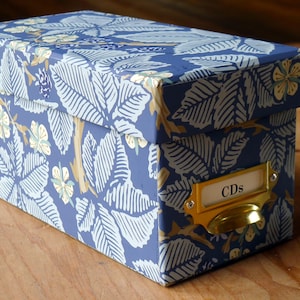 May include: A rectangular storage box with a lid, featuring a blue floral pattern with white and gold accents. The box has a brass label holder that reads "CDs".