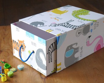 May include: A white gift box with a colorful animal print featuring a tiger, zebra, and flamingo. The box has a blue rope handle and contains small toy accessories. The box is on a wooden surface.
