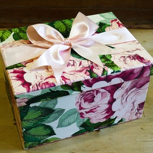 May include: A white gift box with a pink floral pattern and a large pink satin bow. The box is decorated with pink roses and green leaves.