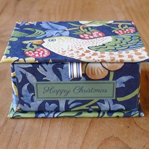 Op de afbeelding: Kleine, decoratieve doos met deksel, met een blauw bloemen- en vogelpatroon. De doos heeft een groen label met de tekst "Happy Christmas". Geplaatst op een houten oppervlak.